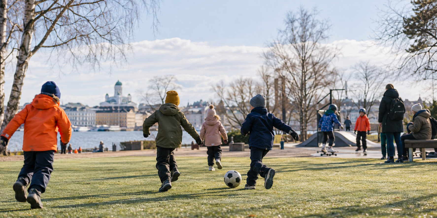 Lapset pelaavat rennosti jalkapalloa Helsingin puistomaisessa ympäristössä aikuisten ollessa lähellä.